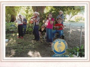 OOTFA District 4 members playing fiddle, guitar and other instruments outdoors under shade trees