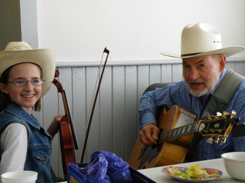 A young girl holding a fiddle smiles next to an elderly man playing guitar, both wearing cowboy hats