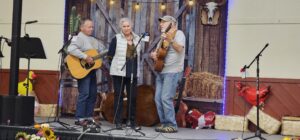 Three OOTFA musicians performing together on stage with guitar, fiddle, and vocals